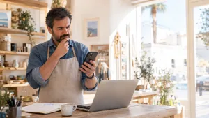 Propietario de un comercio consulta su móvil junto a un portátil, una libreta y una taza de café.